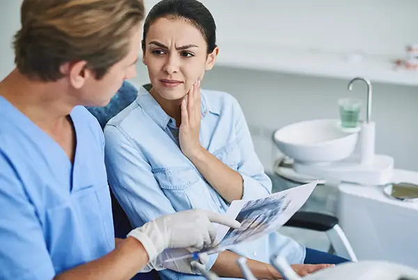 A dentist in scrubs discussing an X-ray with a female patient holding her jaw, indicating TMJ pain or dysfunction.